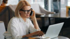 Woman in white shirt and glasses taking a phone call while working on a laptop at a cafe table with a coffee cup beside her.