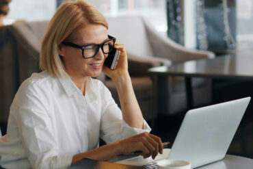 Woman in white shirt and glasses taking a phone call while working on a laptop at a cafe table with a coffee cup beside her.