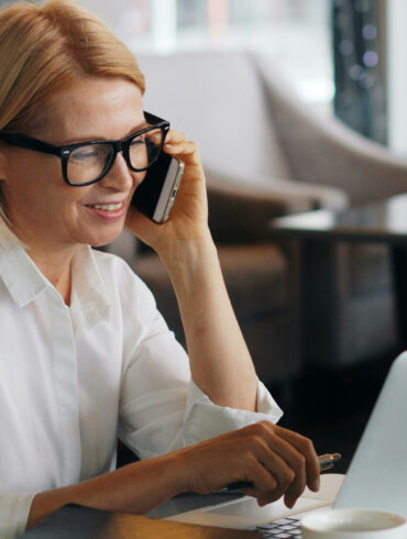 Woman in white shirt and glasses taking a phone call while working on a laptop at a cafe table with a coffee cup beside her.