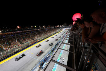 F1 cars lined up on the starting grid at the Singapore Grand Prix night race, with packed grandstands and glowing lanterns overhead.