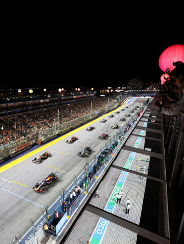 F1 cars lined up on the starting grid at the Singapore Grand Prix night race, with packed grandstands and glowing lanterns overhead.
