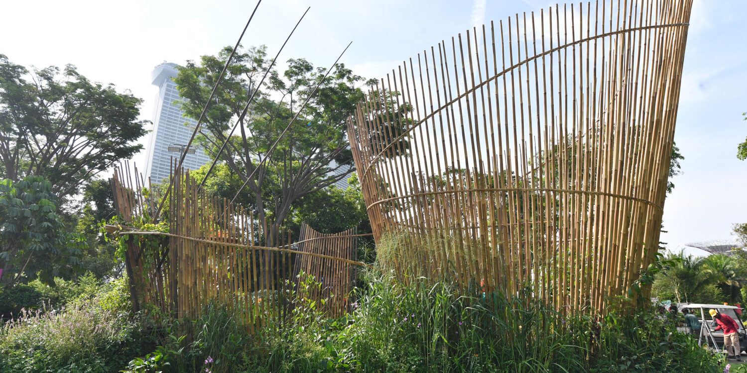 A show garden installation at Singapore Garden Festival featuring curved bamboo screens rising above a dense bed of tropical plants, with Marina Bay Sands visible in the background.