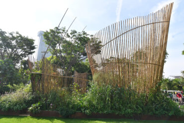A show garden installation at Singapore Garden Festival featuring curved bamboo screens rising above a dense bed of tropical plants, with Marina Bay Sands visible in the background.