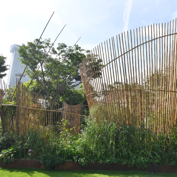 A show garden installation at Singapore Garden Festival featuring curved bamboo screens rising above a dense bed of tropical plants, with Marina Bay Sands visible in the background.
