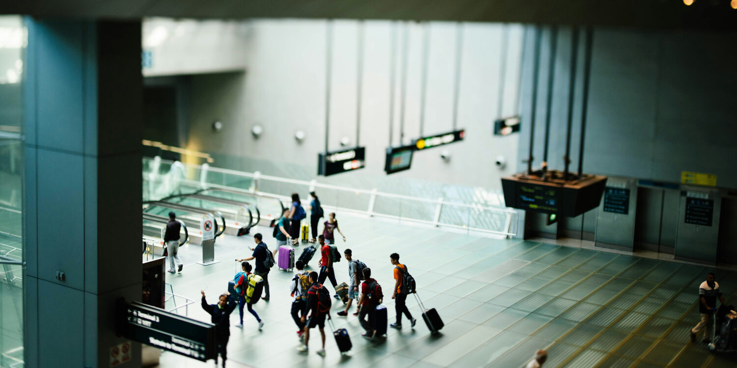 A group of travellers with luggage walks across the polished floor of a Changi Airport terminal, with directional signs to Terminal 3 and the Skytrain visible in the foreground.