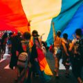 Crowd of people walking under a large rainbow Pride flag at an outdoor event
