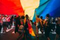 Crowd of people walking under a large rainbow Pride flag at an outdoor event