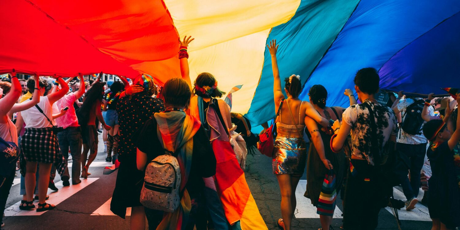 Crowd of people walking under a large rainbow Pride flag at an outdoor event