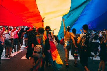 Crowd of people walking under a large rainbow Pride flag at an outdoor event