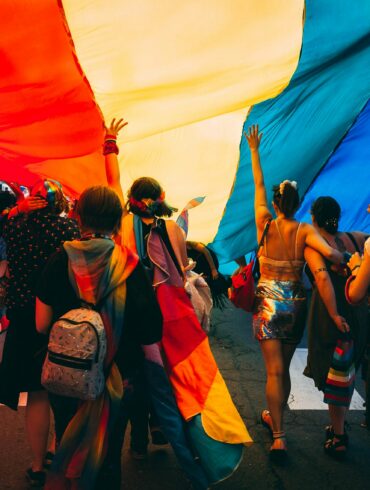 Crowd of people walking under a large rainbow Pride flag at an outdoor event