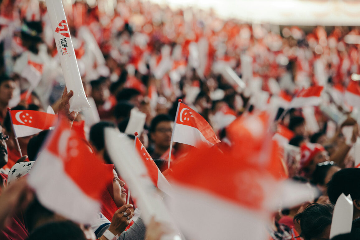 Crowd waving Singapore flags and NDP goodie bag at National Day Parade, with fluorescent light sticks lighting up the stands