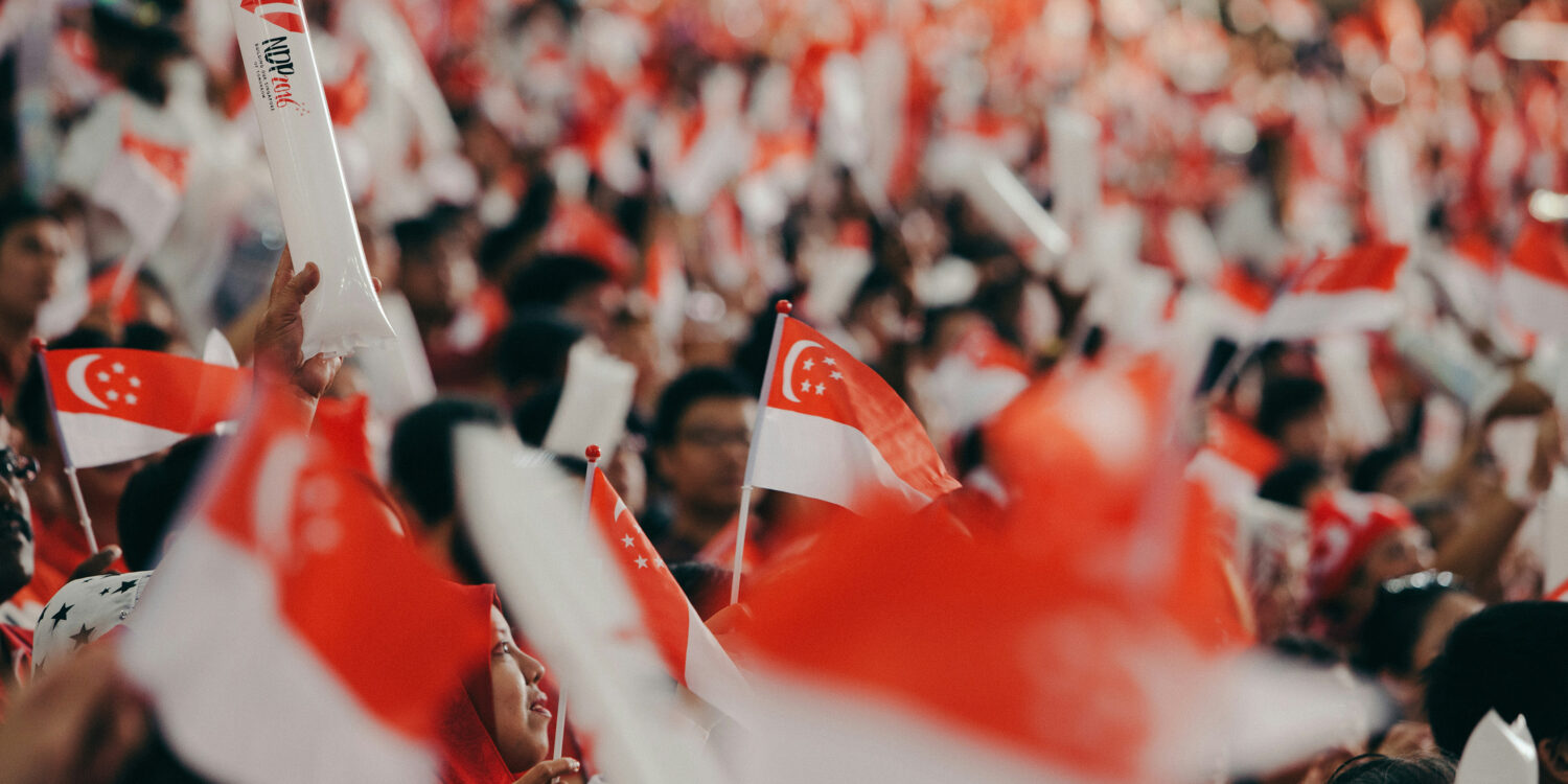 Crowd waving Singapore flags and NDP goodie bag at National Day Parade, with fluorescent light sticks lighting up the stands