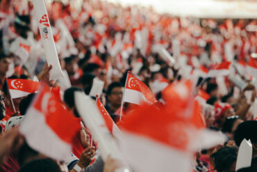 Crowd waving Singapore flags and NDP goodie bag at National Day Parade, with fluorescent light sticks lighting up the stands