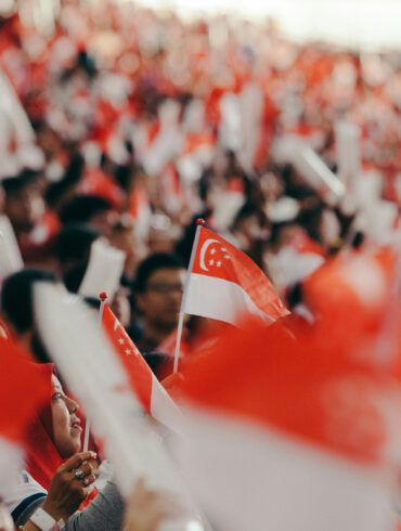 Crowd waving Singapore flags and NDP goodie bag at National Day Parade, with fluorescent light sticks lighting up the stands