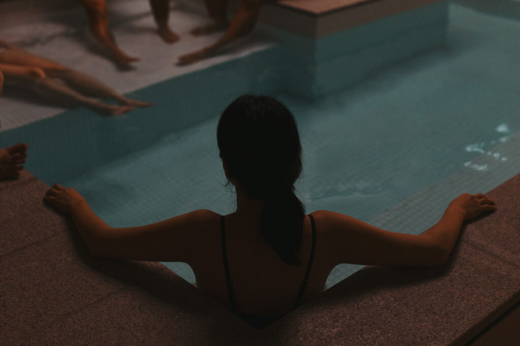 Silhouette of a woman seated at the edge of a bathhouse pool, with other guests visible in the background