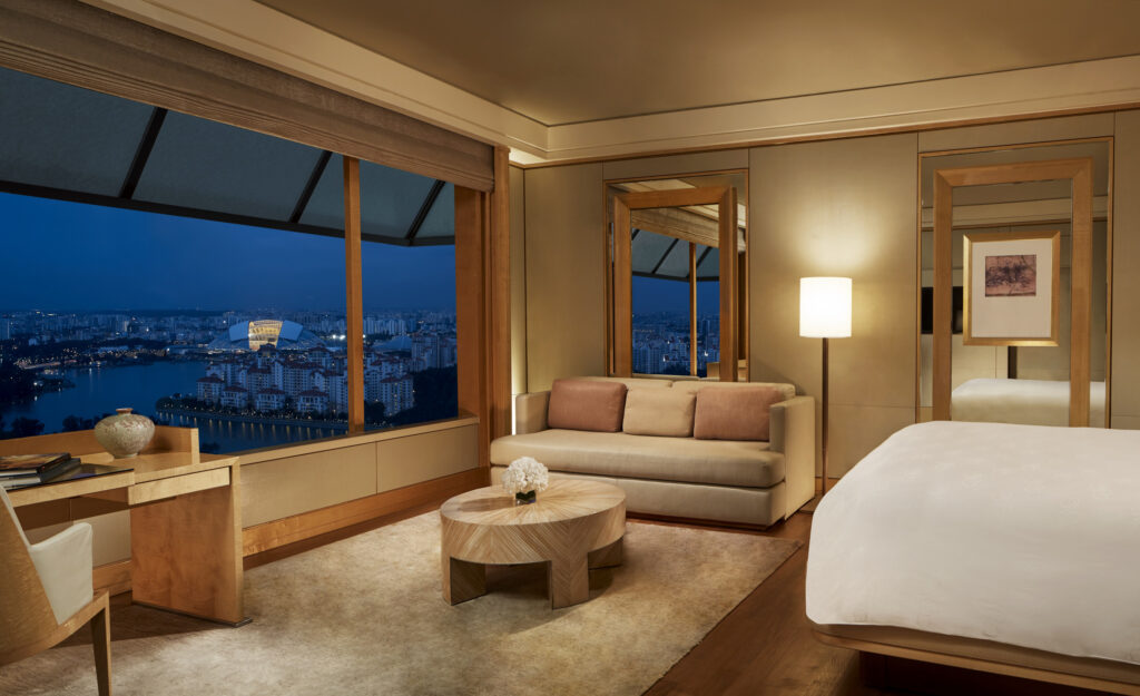 The Ritz-Carlton, Millenia Singapore bedroom with a sofa, floor lamp and round coffee table, with floor-to-ceiling windows overlooking National Stadium and the Kallang Basin skyline at dusk