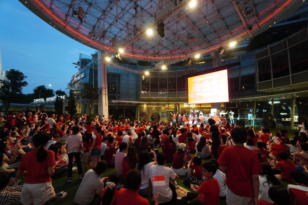 Crowd seated and standing at Capitol Singapore Outdoor Plaza in the evening, waving Singapore flags while facing a stage and screen