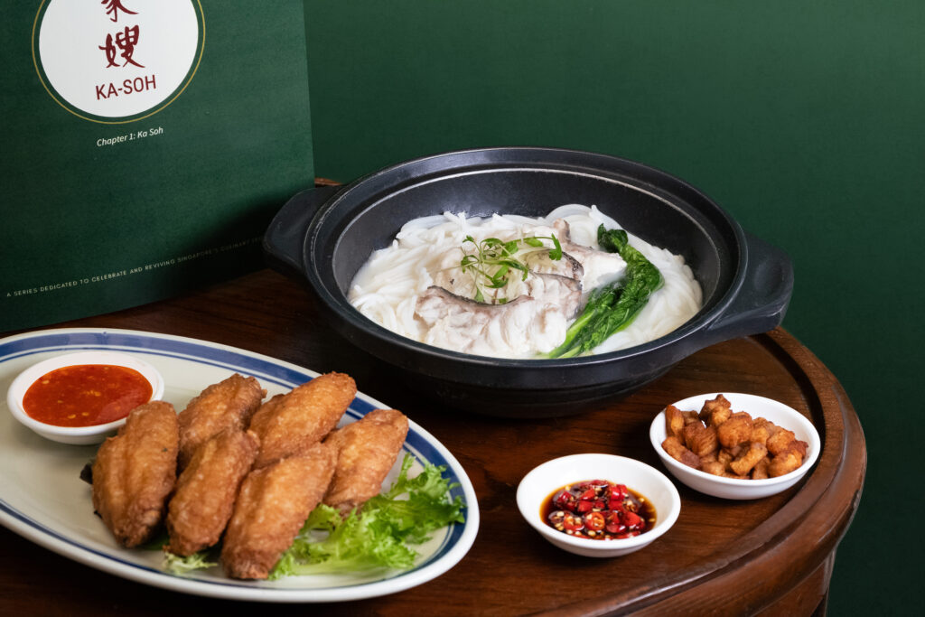 Ka-Soh Signature Fish Noodle Soup in a claypot alongside a plate of Prawn Paste Chicken Mid Wings with dipping sauces, with the Ka-Soh Chapter 1 heritage series signage in the background