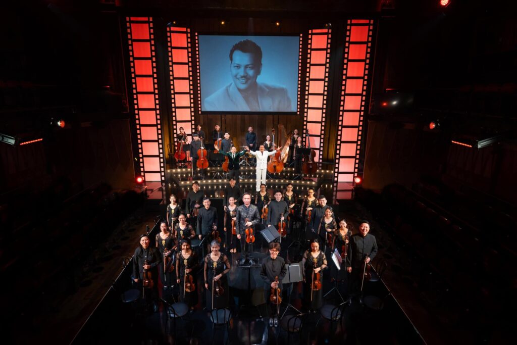 re:mix chamber orchestra and cast take a bow on stage at Wild Rice's Tunggu Sekejap: The P. Ramlee Suite, with a portrait of P. Ramlee projected behind them