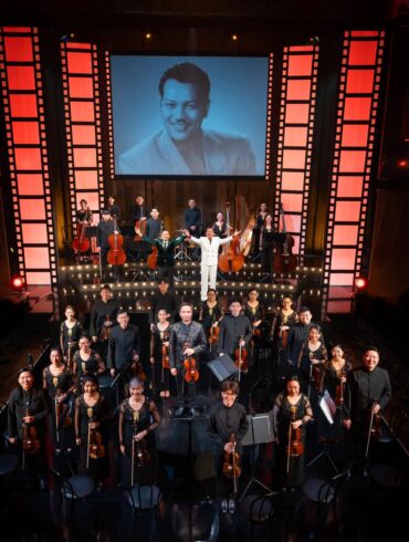 re:mix chamber orchestra and cast take a bow on stage at Wild Rice's Tunggu Sekejap: The P. Ramlee Suite, with a portrait of P. Ramlee projected behind them