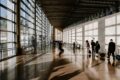 Travellers pulling luggage through a sunlit airport terminal corridor with floor-to-ceiling windows and long shadows across the floor.