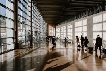 Travellers pulling luggage through a sunlit airport terminal corridor with floor-to-ceiling windows and long shadows across the floor.