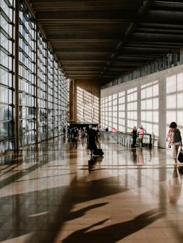Travellers pulling luggage through a sunlit airport terminal corridor with floor-to-ceiling windows and long shadows across the floor.