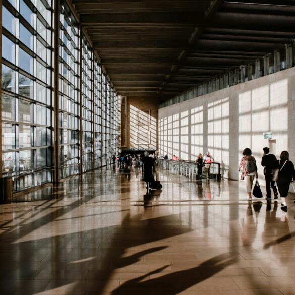 Travellers pulling luggage through a sunlit airport terminal corridor with floor-to-ceiling windows and long shadows across the floor.