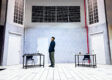 Actor stands alone on the stark, tiled set of Charged by Teater Ekamatra, flanked by two bare tables and chairs under barred windows.