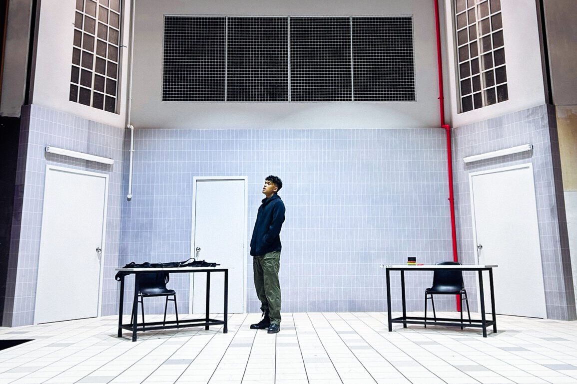 Actor stands alone on the stark, tiled set of Charged by Teater Ekamatra, flanked by two bare tables and chairs under barred windows.