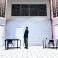 Actor stands alone on the stark, tiled set of Charged by Teater Ekamatra, flanked by two bare tables and chairs under barred windows.