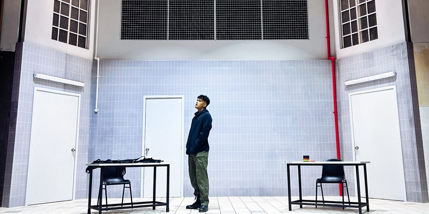 Actor stands alone on the stark, tiled set of Charged by Teater Ekamatra, flanked by two bare tables and chairs under barred windows.