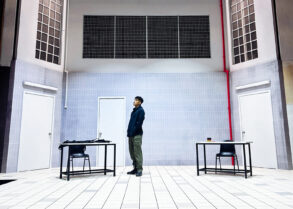 Actor stands alone on the stark, tiled set of Charged by Teater Ekamatra, flanked by two bare tables and chairs under barred windows.