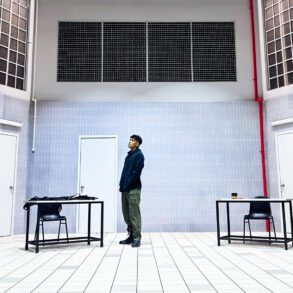 Actor stands alone on the stark, tiled set of Charged by Teater Ekamatra, flanked by two bare tables and chairs under barred windows.