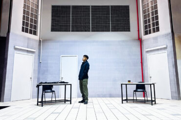 Actor stands alone on the stark, tiled set of Charged by Teater Ekamatra, flanked by two bare tables and chairs under barred windows.