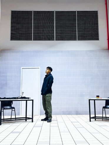 Actor stands alone on the stark, tiled set of Charged by Teater Ekamatra, flanked by two bare tables and chairs under barred windows.