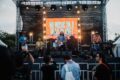 A band performs on an outdoor stage with a large illuminated screen displaying 'NO MUSIC ON A DEAD PLANET' as audience members watch from below.