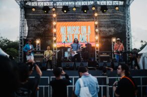 A band performs on an outdoor stage with a large illuminated screen displaying 'NO MUSIC ON A DEAD PLANET' as audience members watch from below.