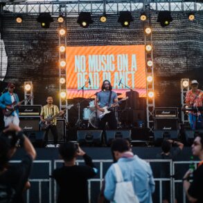 A band performs on an outdoor stage with a large illuminated screen displaying 'NO MUSIC ON A DEAD PLANET' as audience members watch from below.