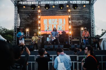 A band performs on an outdoor stage with a large illuminated screen displaying 'NO MUSIC ON A DEAD PLANET' as audience members watch from below.