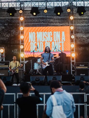A band performs on an outdoor stage with a large illuminated screen displaying 'NO MUSIC ON A DEAD PLANET' as audience members watch from below.