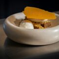 A plated dessert of spiced cake with crumble, quenelles of cream and ice cream, and a translucent orange tuile, served in a rounded stone bowl.