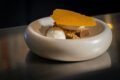 A plated dessert of spiced cake with crumble, quenelles of cream and ice cream, and a translucent orange tuile, served in a rounded stone bowl.