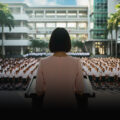 School principal in pink dress addresses uniformed students at assembly, key art for The Radicalisation of Mrs. Mary Lim-Rodrigues at Wild Rice