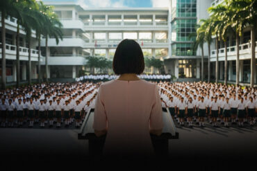 School principal in pink dress addresses uniformed students at assembly, key art for The Radicalisation of Mrs. Mary Lim-Rodrigues at Wild Rice