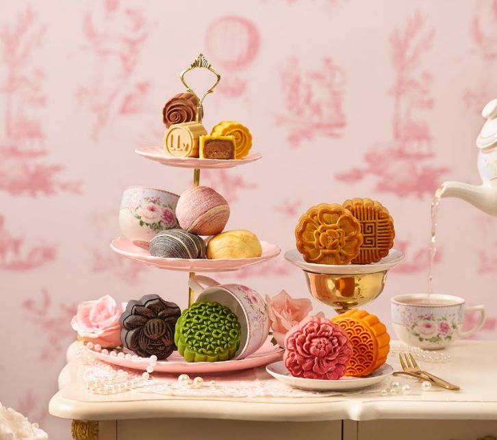 An assortment of decorative mooncakes in various colours and patterns displayed on a pink three-tier stand and plates.