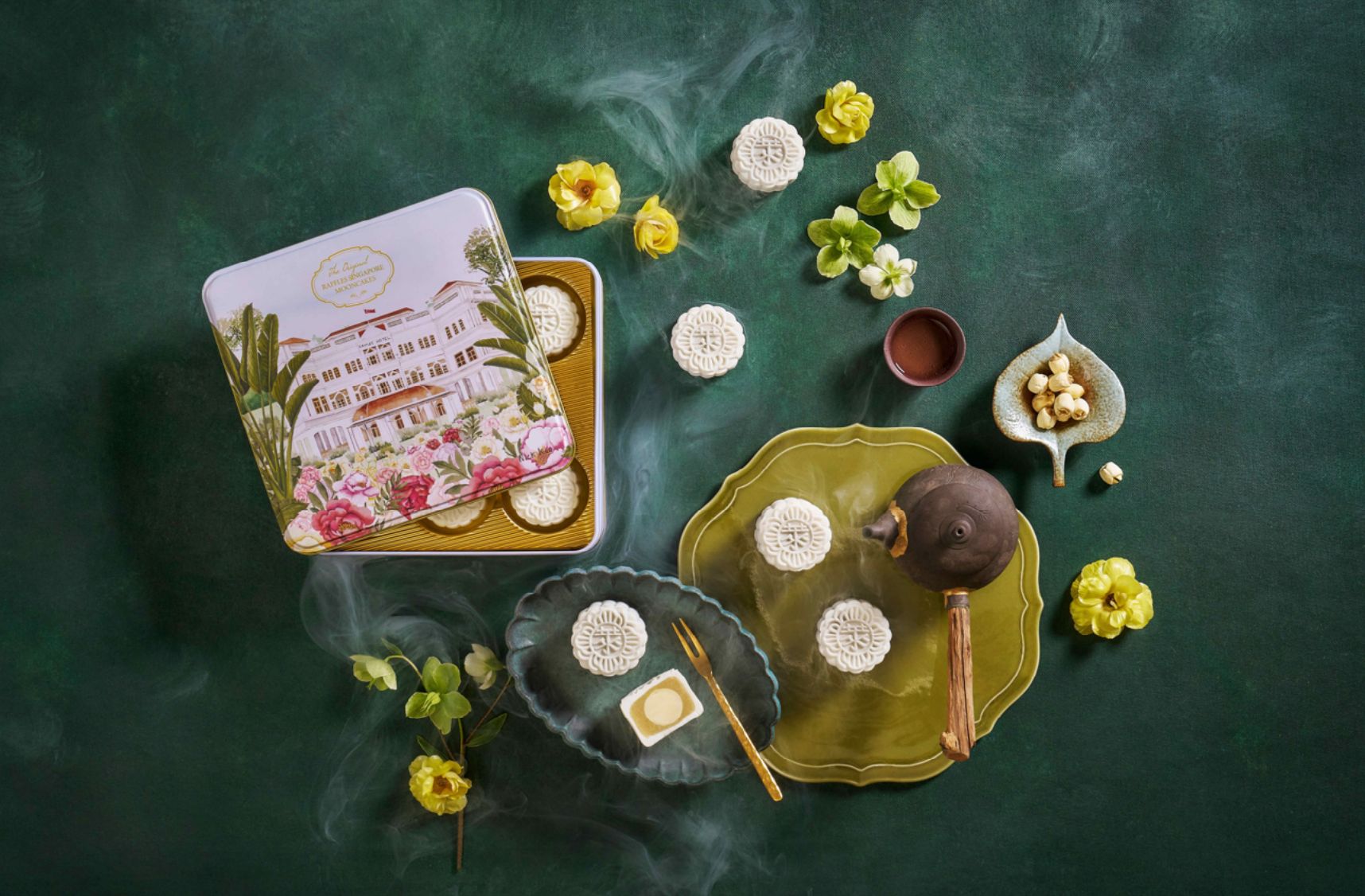 An ornate gift tin and traditional white mooncakes are arranged with decorative plates, flowers and a chocolate-brown teapot on a green marble surface.
