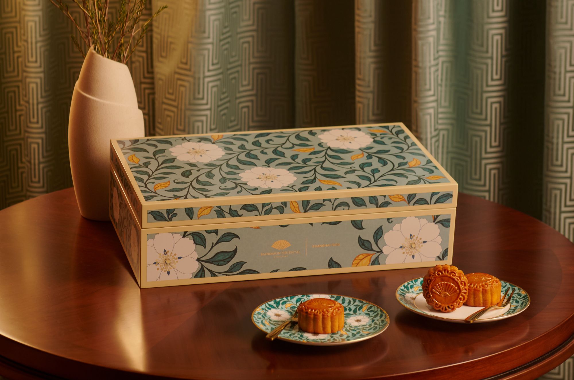 A decorative mooncake gift box with floral patterns sits on a wooden table alongside matching plates holding traditional mooncakes.