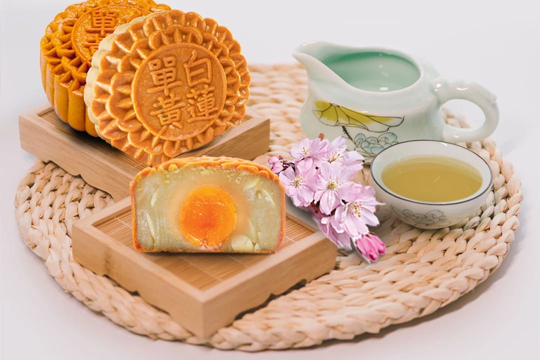 Golden-brown mooncakes with decorative patterns are displayed on wooden boards alongside a halved mooncake revealing its salted egg yolk filling, with tea and cherry blossoms arranged on a woven mat.