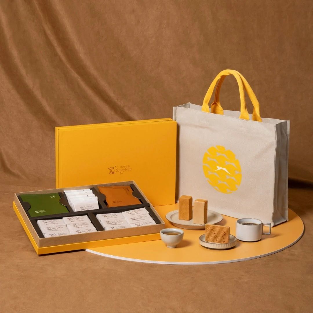 A gift set displays yellow-packaged pastry boxes alongside a white tote bag with a pineapple motif, arranged with tea cups and baked goods.